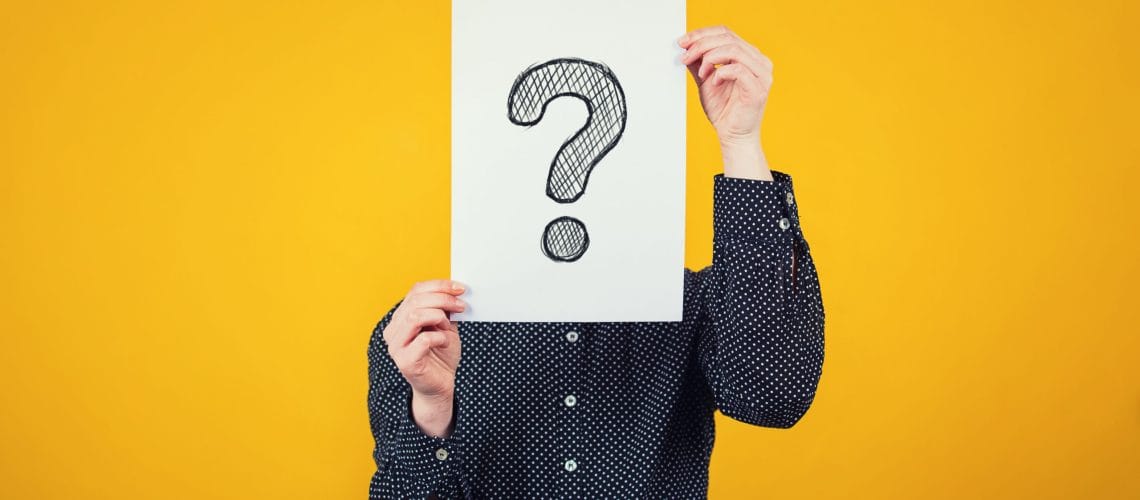 Businesswoman covering face using a white paper sheet with drawn question mark, like a mask, for hiding her identity. Isolated on yellow wall background